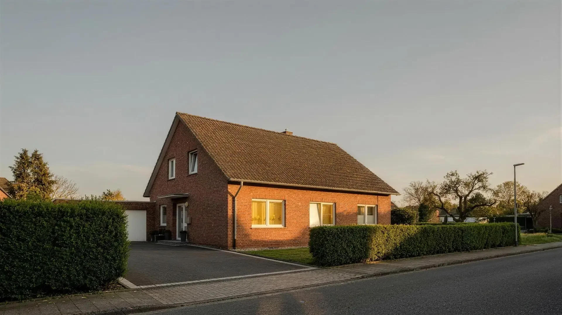 Ein Einfamilienhaus mit Klinkerfassade und hellen Fenstern am Abend hinter einer Hecke (Stimmungsbild)